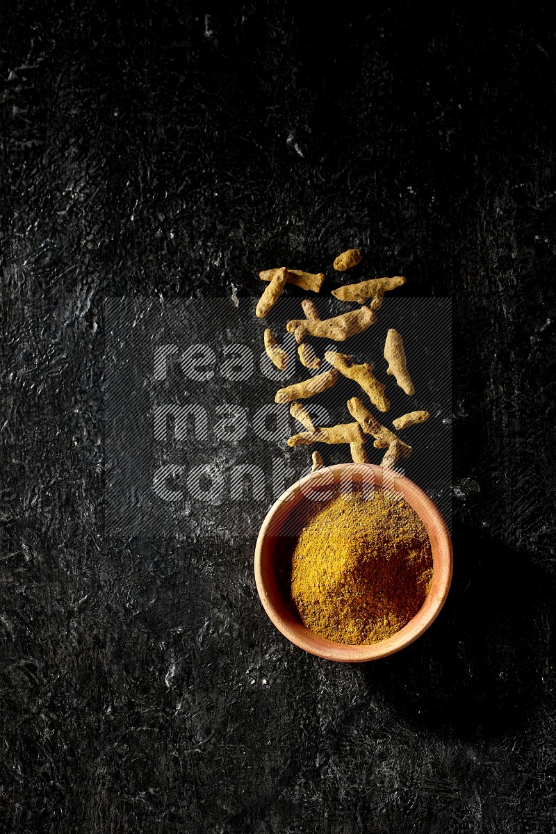 A wooden bowl full of turmeric powder with dried turmeric fingers on textured black flooring