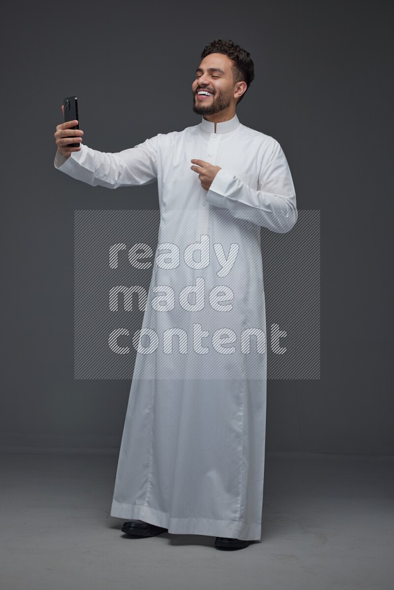 A Saudi man wearing Thobe and taking selfie with his phone making different poses eye level on a gray background