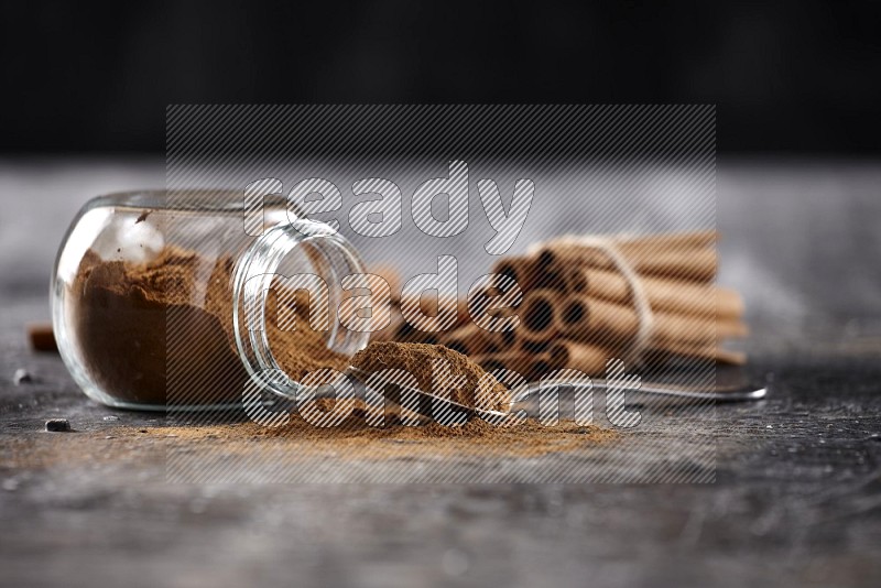 Herbal glass jar full cinnamon powder flipped and a metal spoon full of powder, cinnamon sticks stacked and bounded in the back on textured black background in different angles