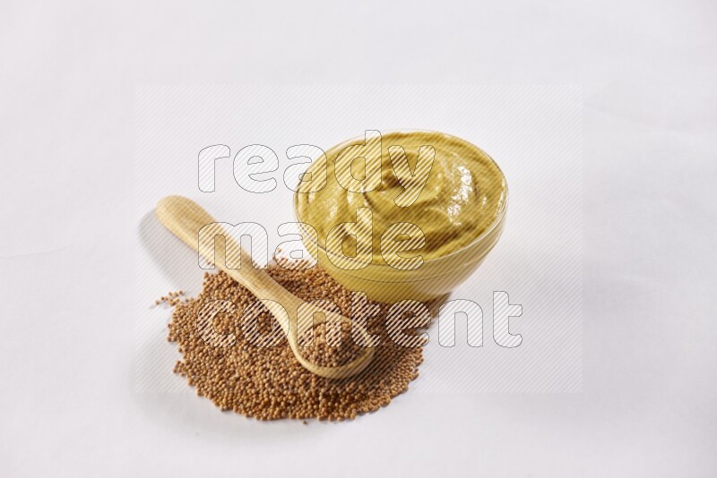 A glass bowl full of mustard paste with some of mustard seeds underneath it and a wooden spoon on white flooring
