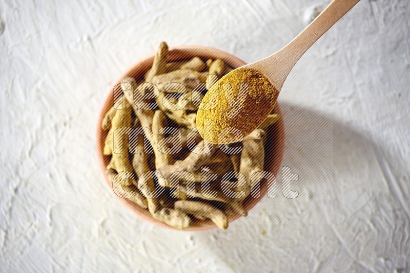 A wooden spoon full of turmeric powder above a wooden bowl full of dried turmeric whole fingers on a textured white flooring