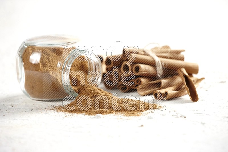 Flipped herbs glass jar full of cinnamon powder and cinnamon sticks in the back on a textured white background