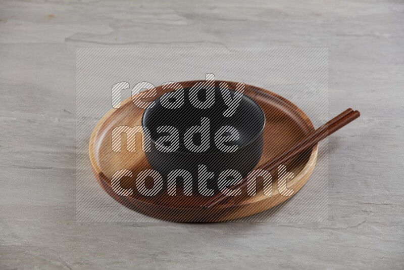 black ceramic round bowl on top of brown wood round plate and wood chopsticks, on grey textured countertop