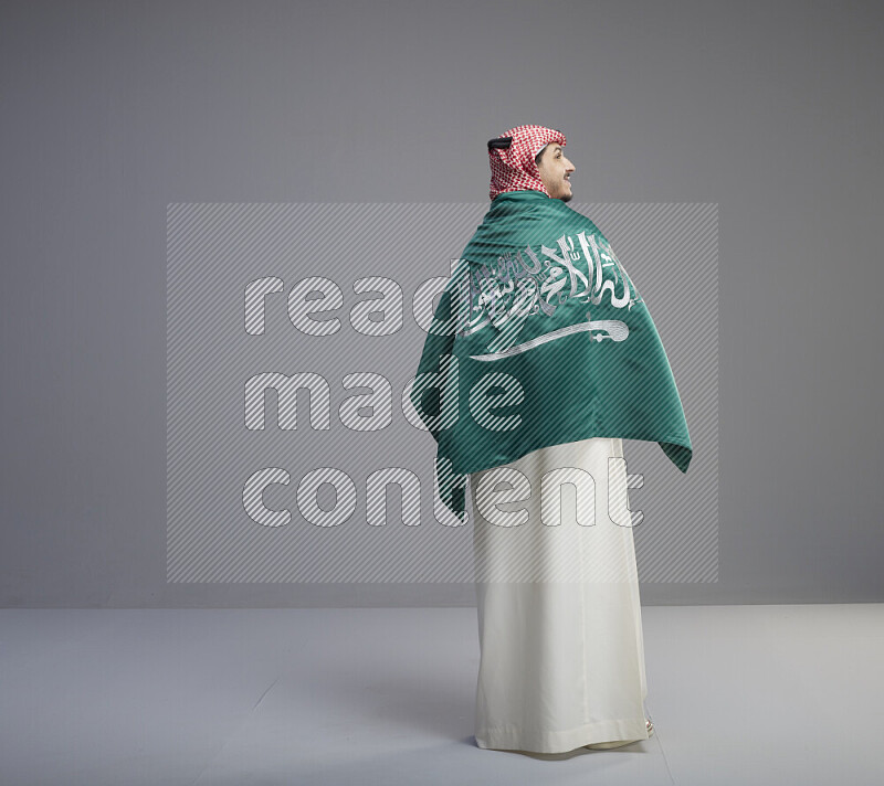 A Saudi man standing wearing thob and red shomag wrapping big saudi flag on gray background