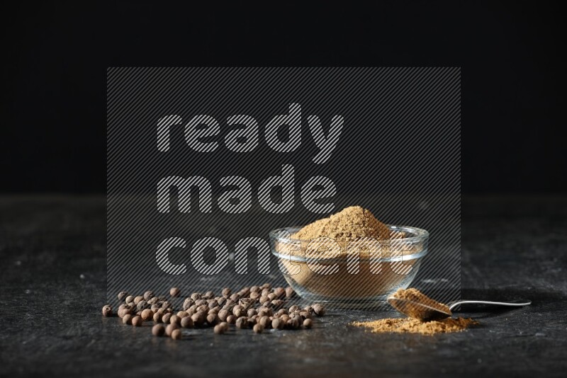 A glass bowl and metal spoon full of allspice powder and whole balls spreaded on a black flooring