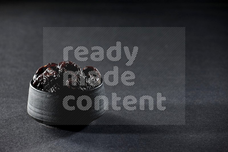 A black pottery bowl full of dried plums on a black background in different angles