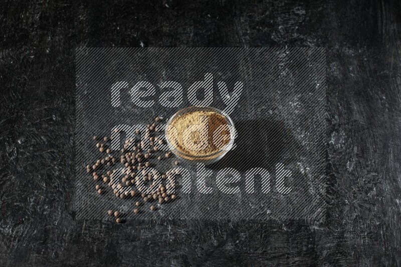 A glass bowl full of allspice powder and whole balls spreaded on a black flooring