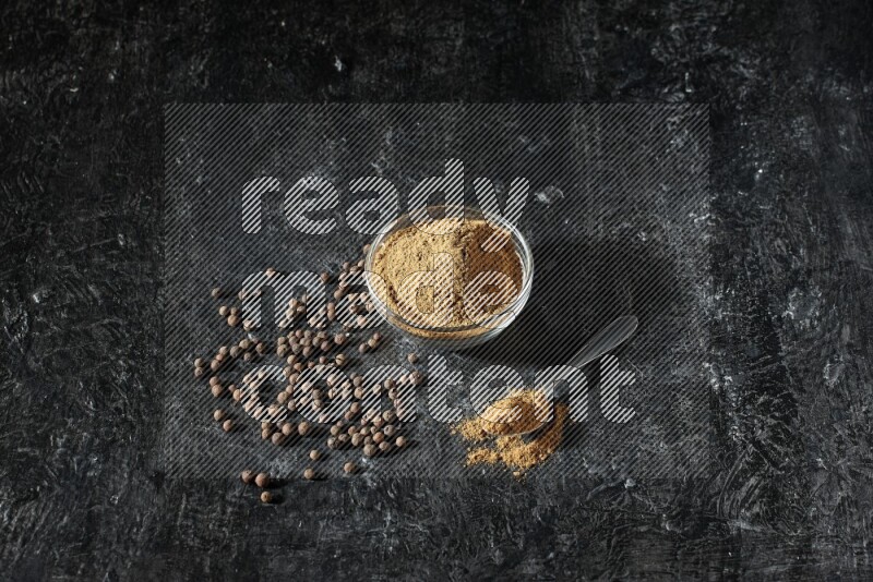 A glass bowl and metal spoon full of allspice powder and whole balls spreaded on a black flooring