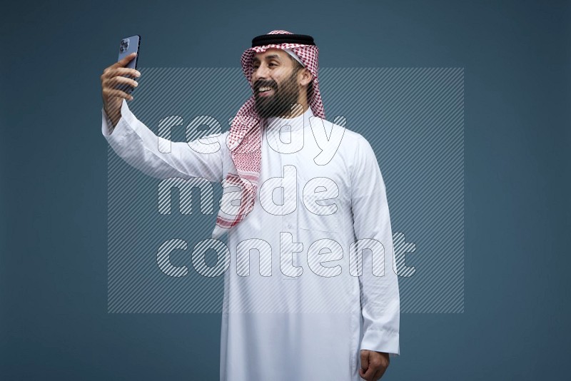 A man Taking Selfie in a blue background wearing Saudi Thob with Shomag