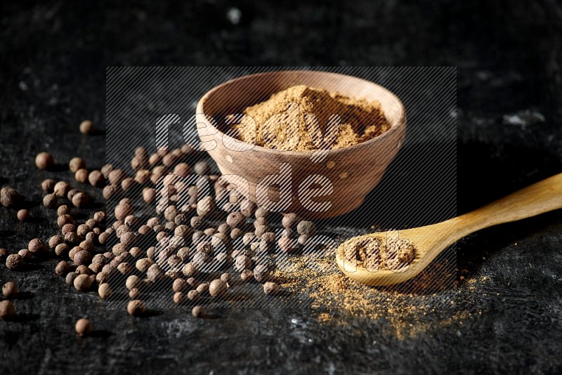 A wooden bowl and spoon full of allspice powder and whole balls spreaded on a black flooring