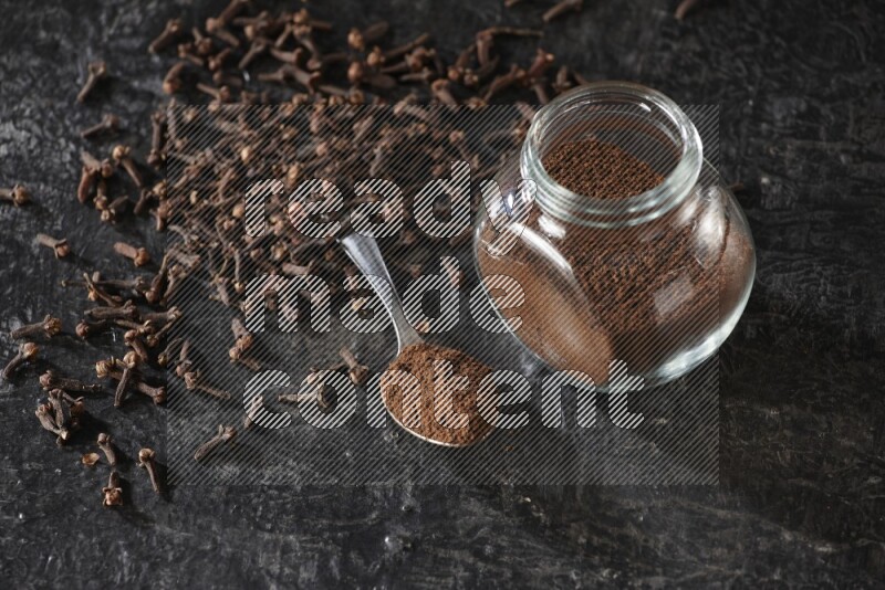 A glass spice jar and a metal spoon full of cloves powder with cloves spread on textured black flooring