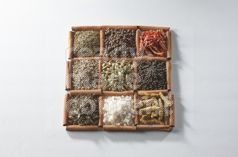 9 مربعات من أعواد القرفة ممتلئة بالهيل في المنتصف محاطة بأصابع الكركم والملح والريحان المجفف والفلفل الأبيض والكمون وبهار الفلفل الحلو والفلفل الحار المجفف والفلفل الأسود على أرضية بيضاء