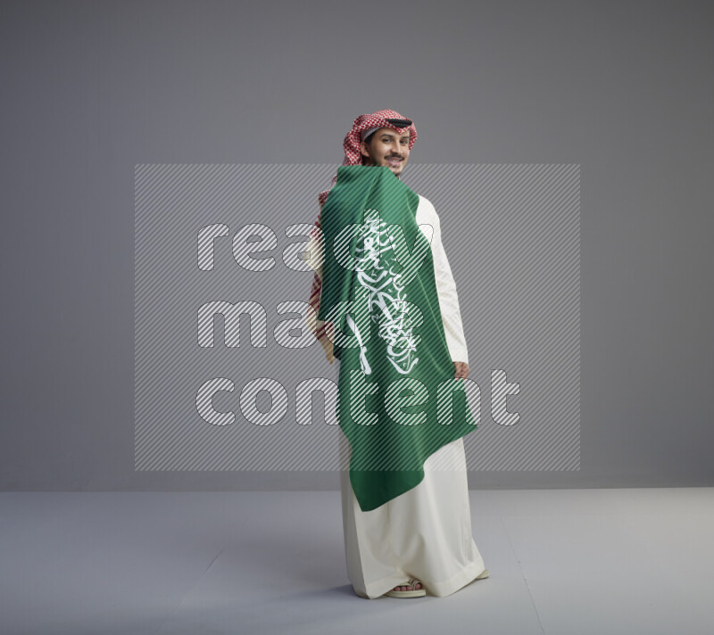 A saudi man standing wearing thob and red shomag holding big saudi flag on gray background