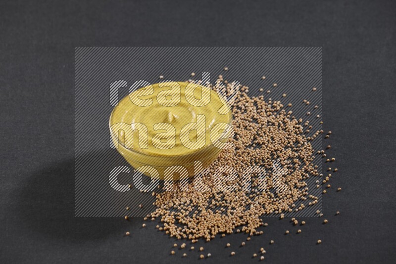 A glass bowl full of mustard paste with mustard seeds underneath on black flooring