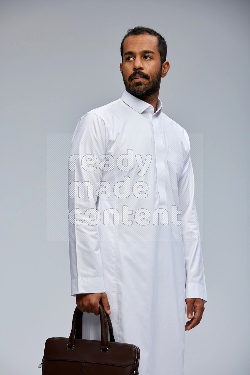 Saudi man wearing thob standing holding suitcase on gray background