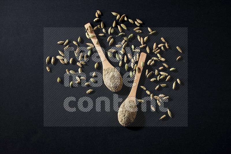 2 Wooden spoons full of cardamom powder and cardamom seeds spreaded on black flooring