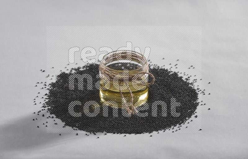 A glass jar full of black seeds oil surrounded by the seeds on a white flooring