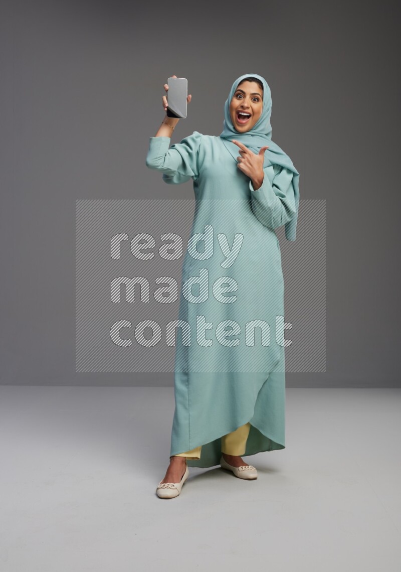Saudi Woman wearing Abaya standing showing phone to camera on Gray background