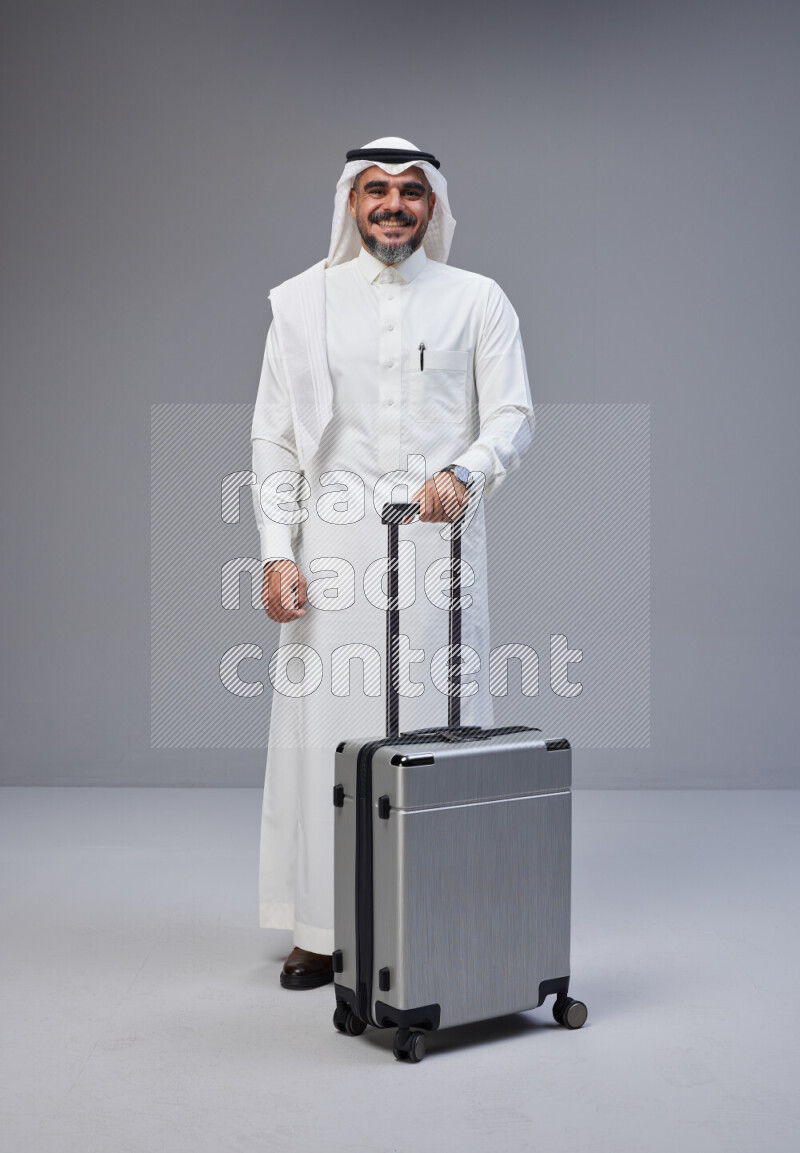 Saudi man wearing Thob and white Shomag standing holding Travel bag on Gray background