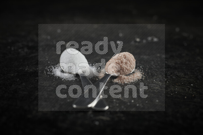 2 metal spoons filled with white salt and pink himalayan salt on black background