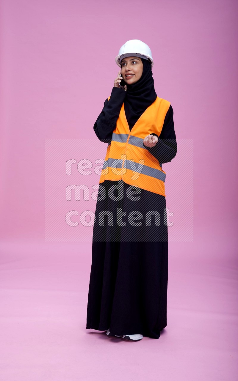 Saudi woman wearing Abaya with engineer vest and helmet standing talking on phone on pink background