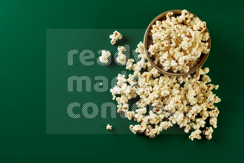 An off white ceramic bowl full of popcorn with popcorn beside it on a green background in a top view shot