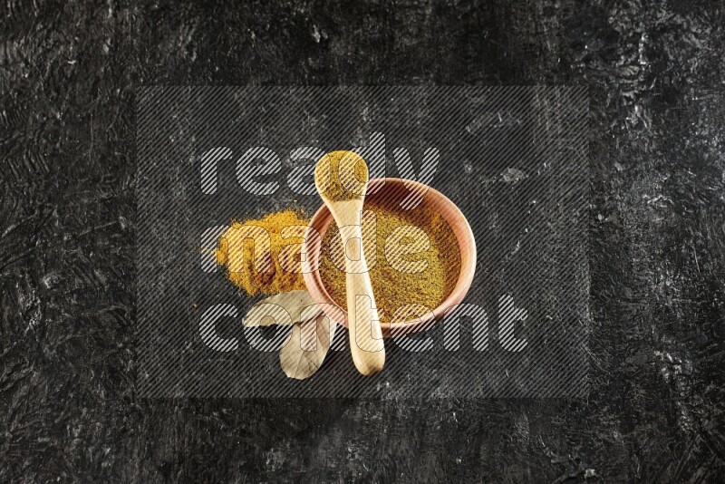 A wooden bowl and a wooden spoon full of turmeric powder on textured black flooring