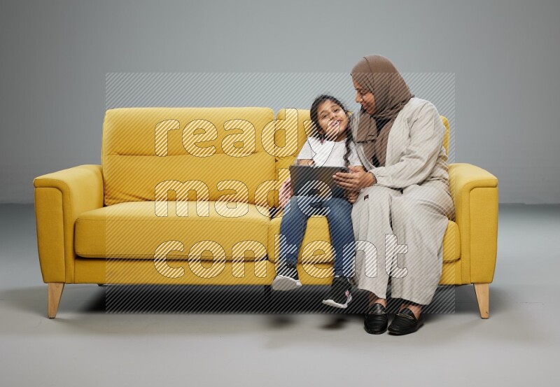 A girl with her mother on yellow sofa and watching on iPad on gray background