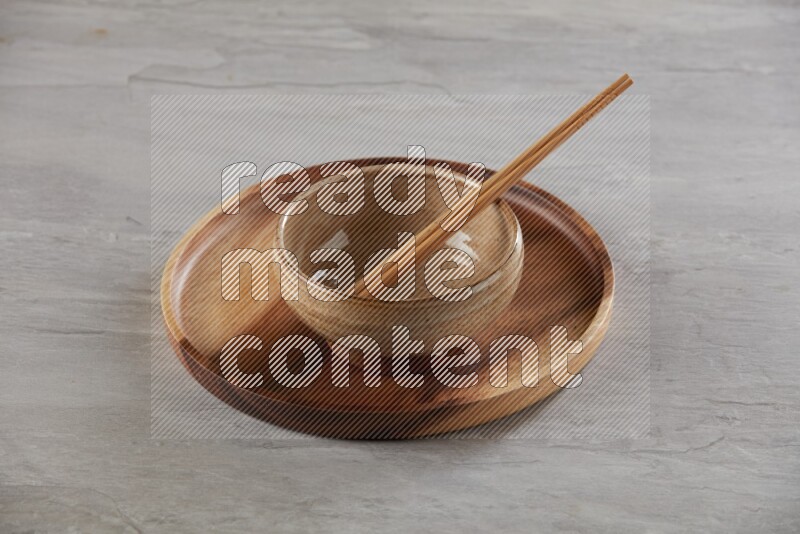multi color pottery round bowl on top of brown wood round plate and wood chopsticks, on grey textured countertop