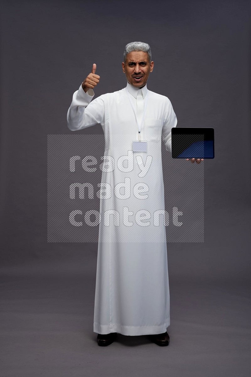 Saudi man wearing thob with neck strap employee badge standing showing tablet to camera on gray background