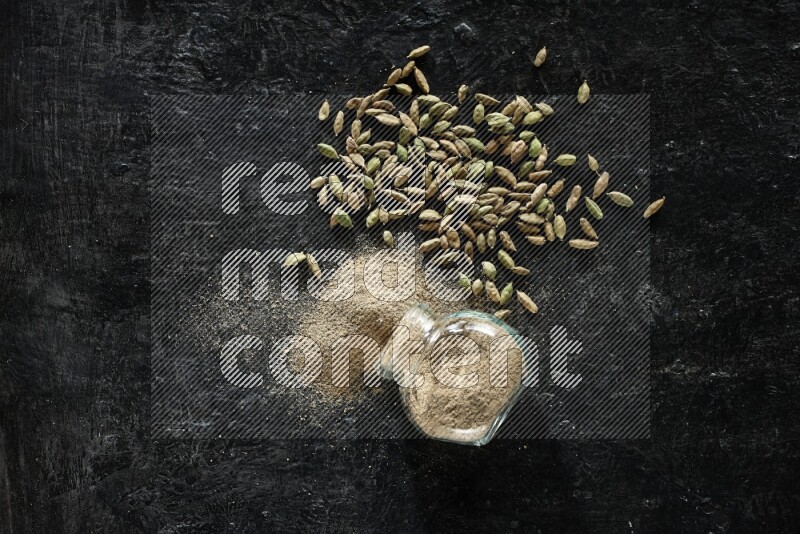 وعاء زجاجي للتوابل مقلوب وممتلئ ببودرة الهيل والبودرة تناثرت منه مع حبوب الهيل المتناثرة على خلفية سوداء