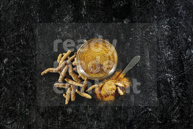 A glass bowl and a metal spoon full of turmeric powder and dried whole fingers next of them on textured black flooring