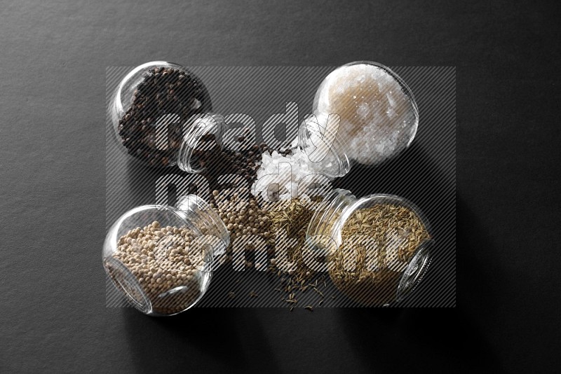 4 glass spice jars full of salt, black peppers, white peppers and cumin on black flooring