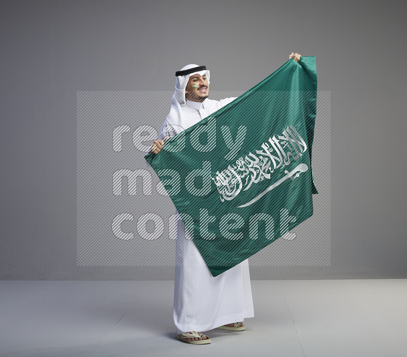 رجل سعودي يرتدي ثوب ابيض وشماغ يحمل العلم السعودي