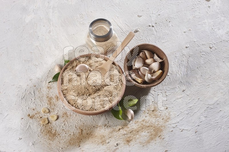 A wooden bowl with a spoon in it and glass spice jar, all full of garlic powder and a wooden bowl full of garlic cloves on a white flooring