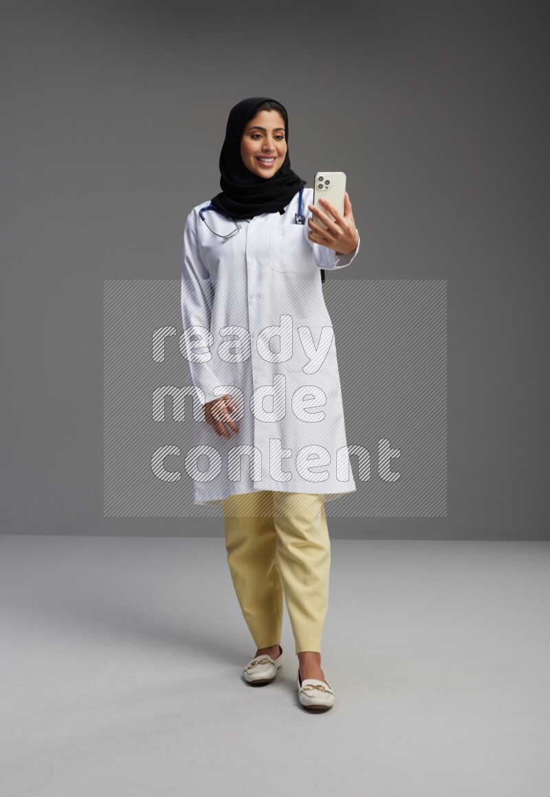 Saudi woman wearing lab coat with stethoscope standing taking selfie on Gray background