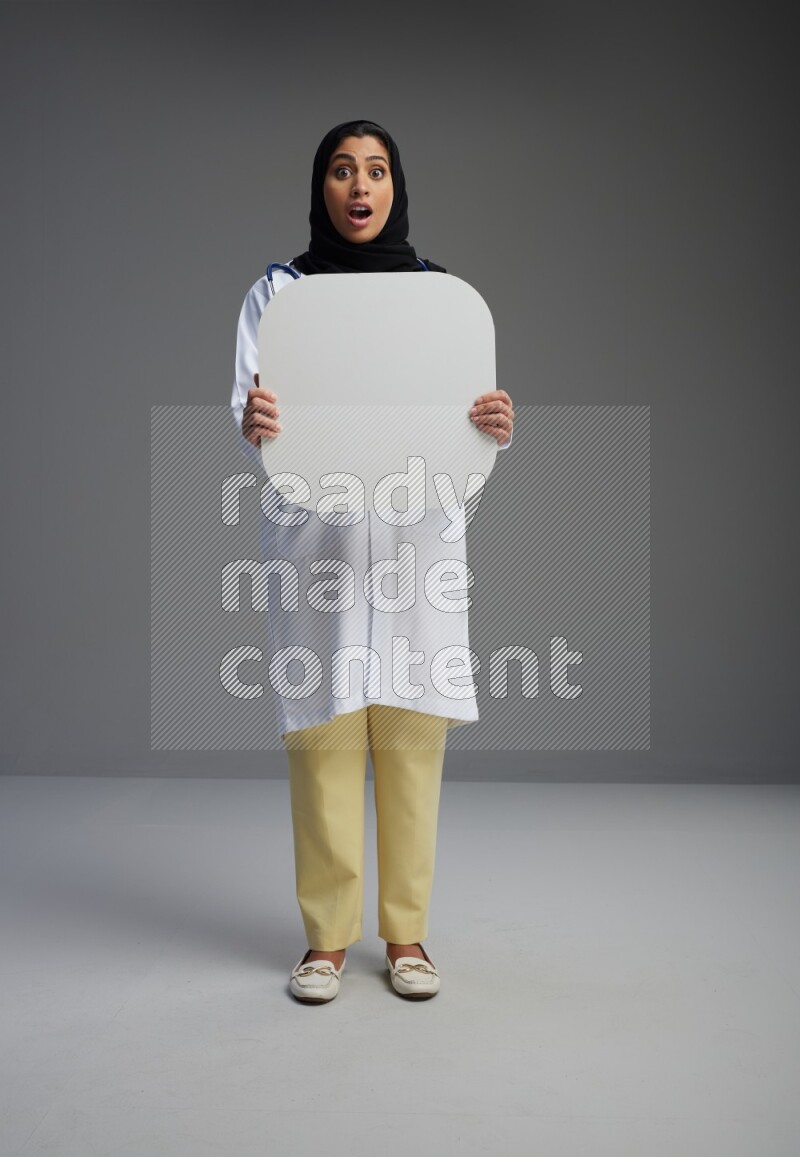 Saudi woman wearing lab coat with stethoscope standing holding social media sign on Gray background