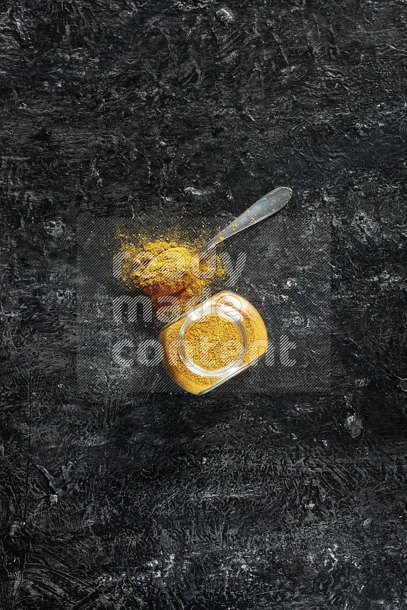 A glass spice jar and metal spoon full of turmeric powder on textured black flooring
