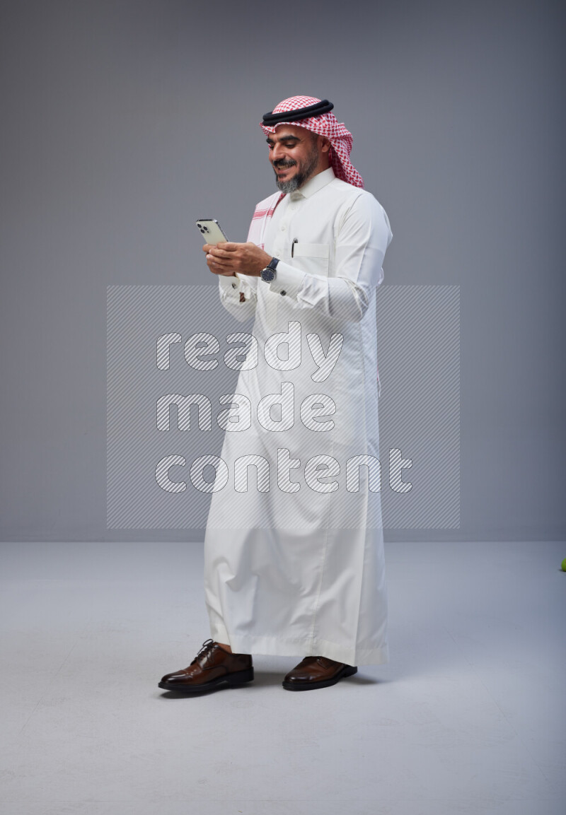 Saudi man Wearing Thob and red Shomag standing texting on phone on Gray background