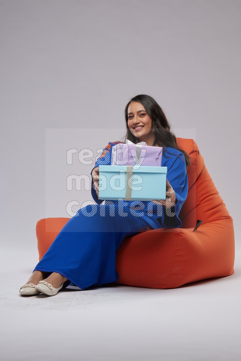 A woman sitting on an orange beanbag wearing Jalabeya holding a gift box