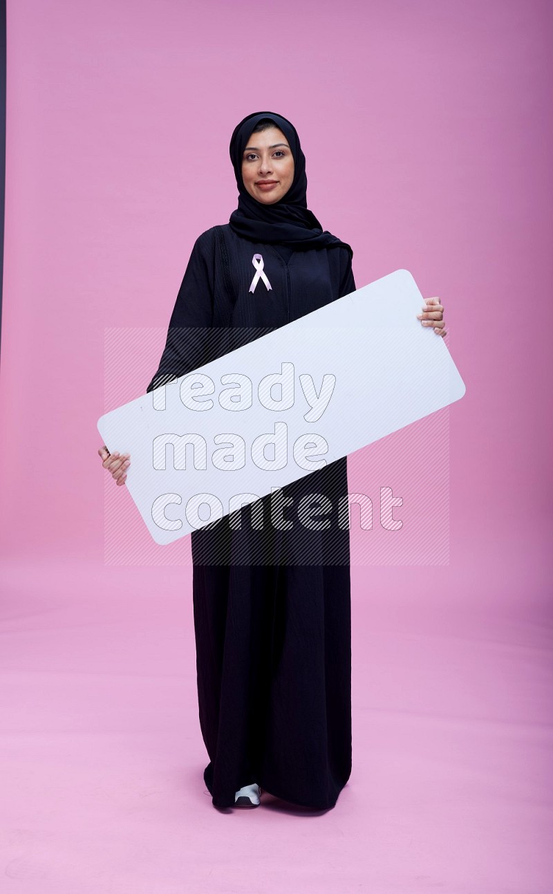 Saudi woman wearing pink ribbon on Abaya standing holding board on pink background