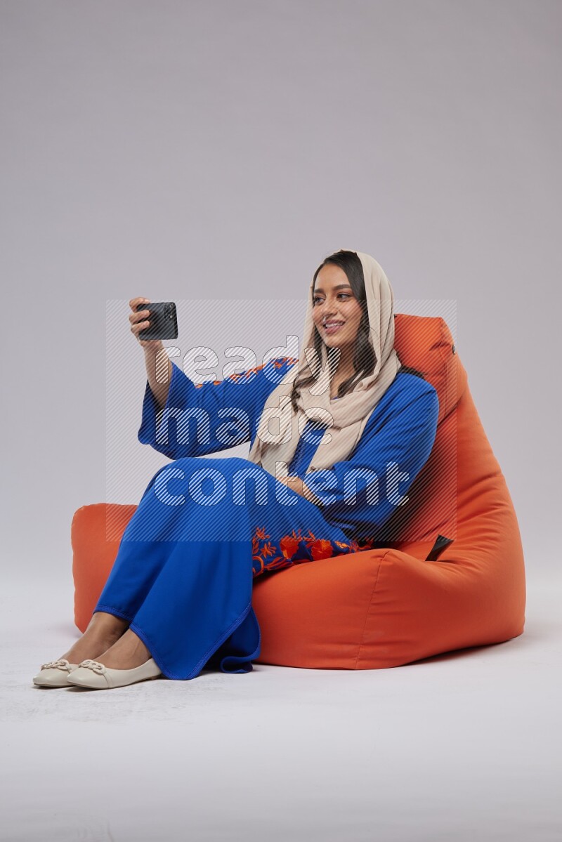 A Saudi woman wearing jalabiya sitting on an orange beanbag and taking selfie