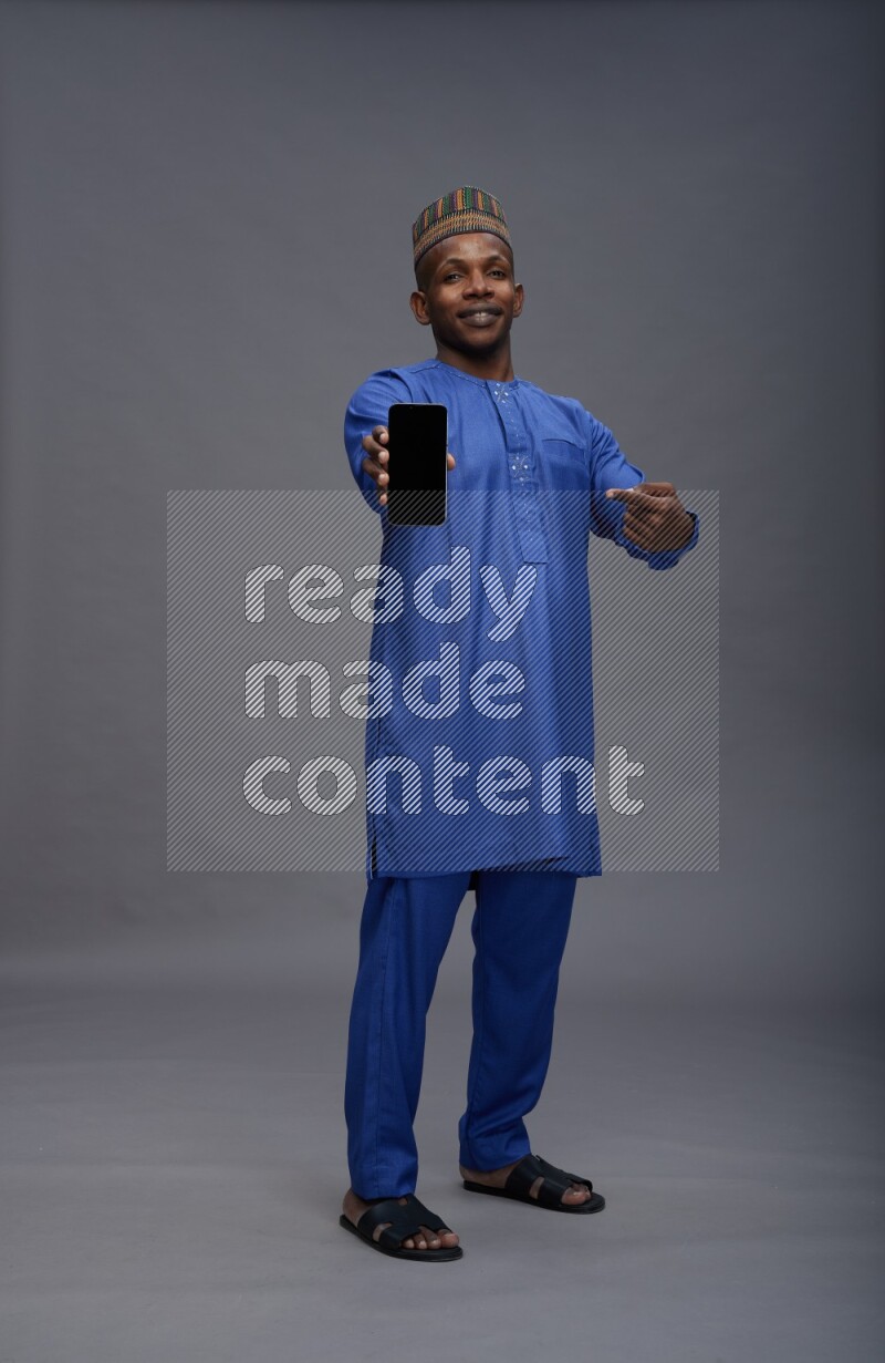 Man wearing Nigerian outfit standing showing phone to camera on gray background