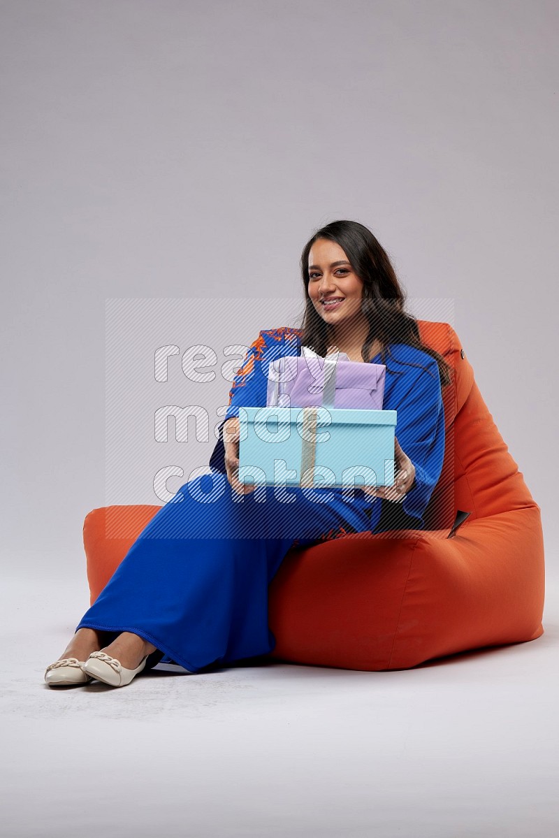 A woman sitting on an orange beanbag wearing Jalabeya holding a gift box