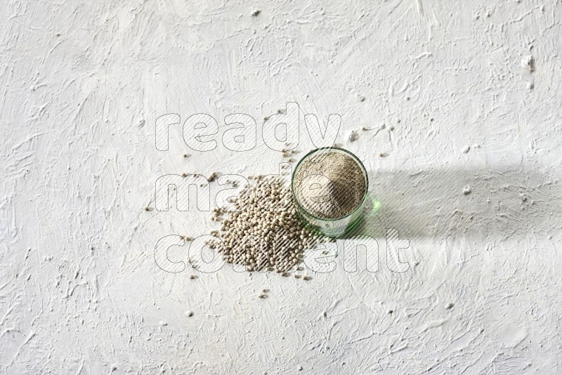 A green glass cup full of white pepper powder with white pepper beads on textured white flooring