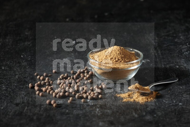 A glass bowl and metal spoon full of allspice powder and whole balls spreaded on a black flooring
