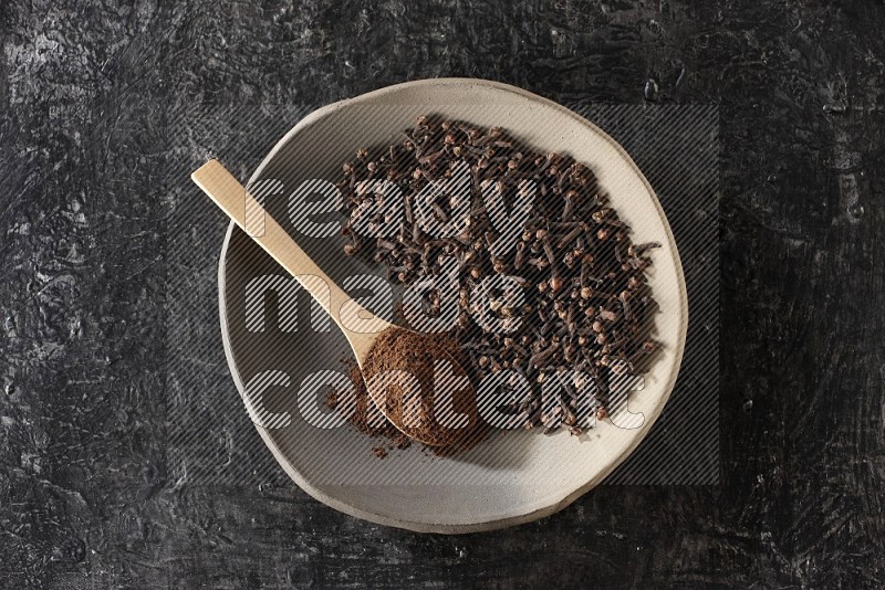 A Pottery plate full of cloves and a wooden spoon full of cloves powder on it on a textured black background