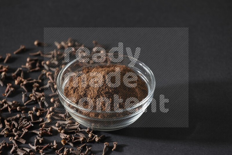 A glass bowl full of cloves powder with cloves grains spread on black flooring