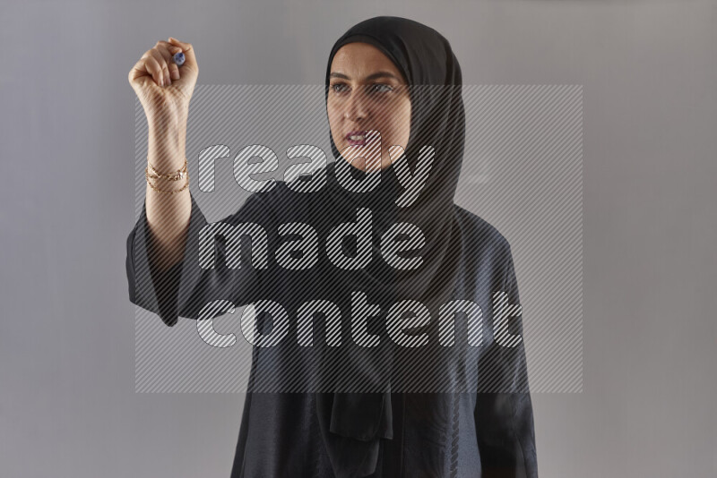 Woman in black abaya and hijab holding a marker pen to write on a transparent board with different reactions and expressions on grey background