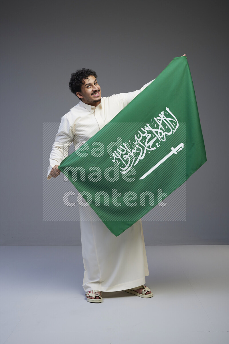 A saudi man standing wearing thob holding big saudi flag on gray background